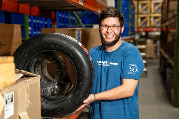 PJi warehouse employee smiling while holding an aircraft tire in the storage area with shelves and boxes in the background.