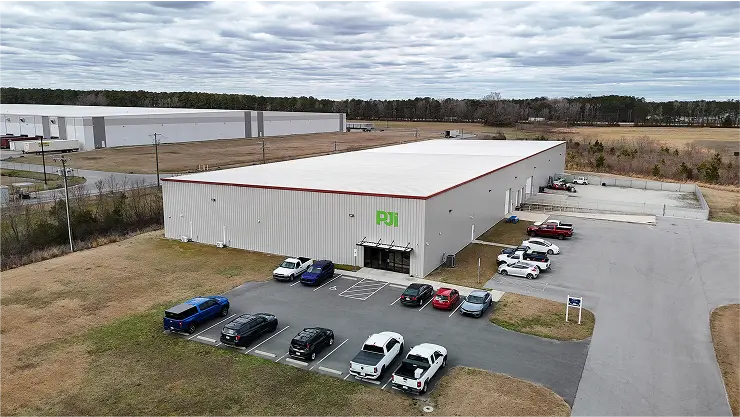 Aerial view of PJiâs metal industrial facility with parking lot and surrounding fields under a cloudy sky.