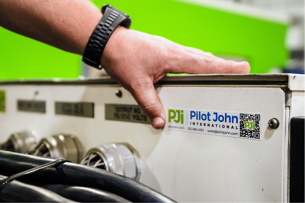 Close-up of a PJi Pilot John International unit with power cables and a technician's hand resting on the equipment.