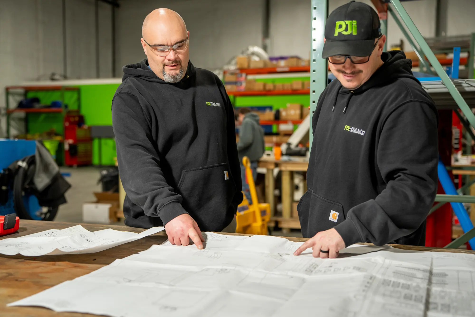 Two PJi team members wearing branded hoodies review large technical drawings together in a workshop filled with tools and equipment.