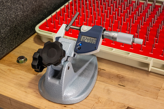 Digital micrometer mounted on a stand with precision gauge pins in the background.