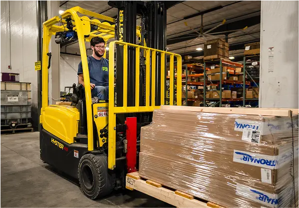 PJi employee operating a yellow Hyster forklift carrying a pallet of boxed Tronair equipment inside the warehouse.