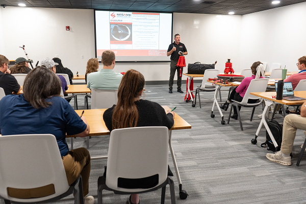 Instructor giving a presentation to a classroom of PJi team members seated at desks during a training session with a slideshow projected on the screen.