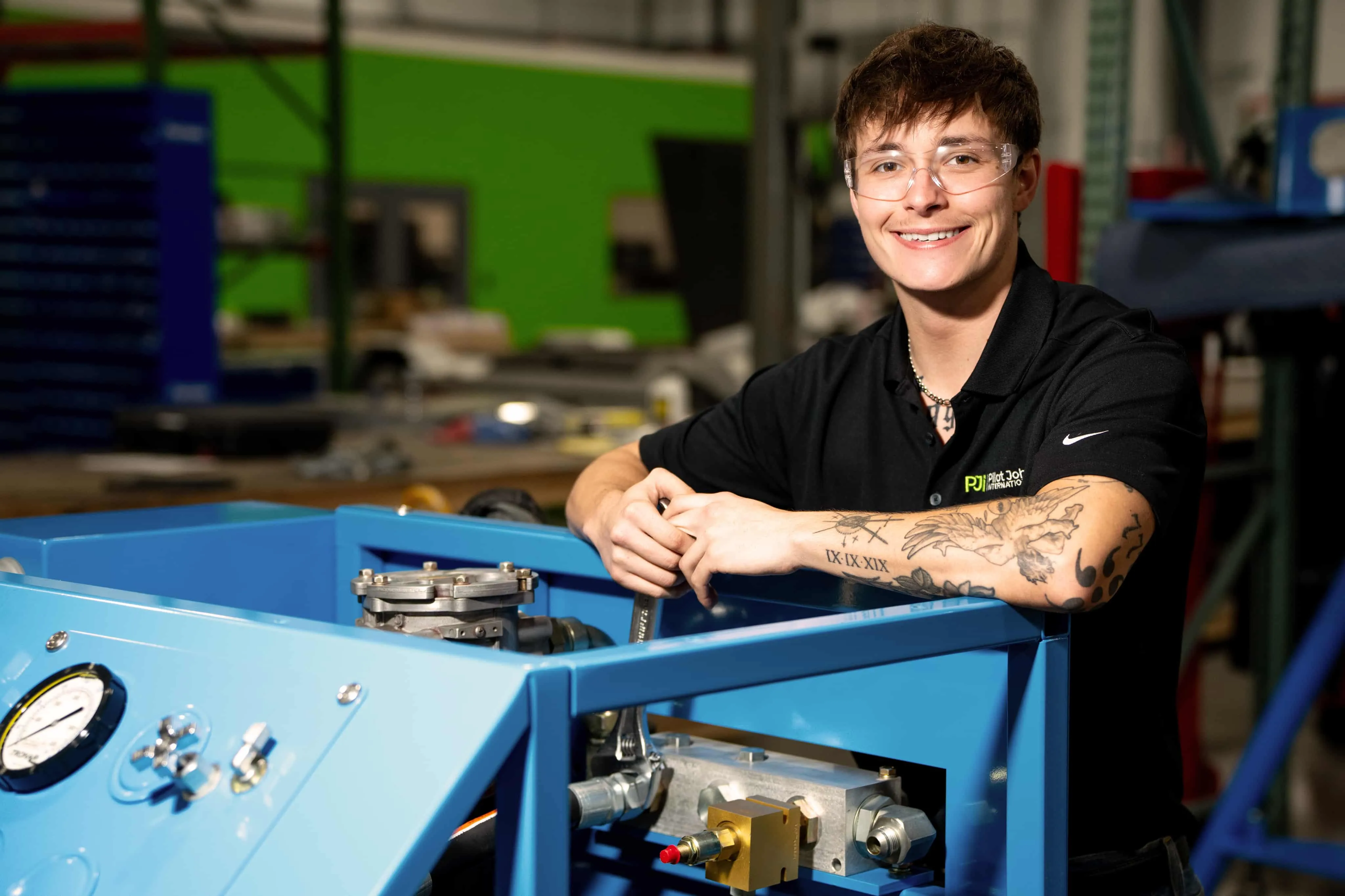 PJi technician smiling while working on blue aviation equipment in the workshop, wearing safety glasses and a black PJi shirt.