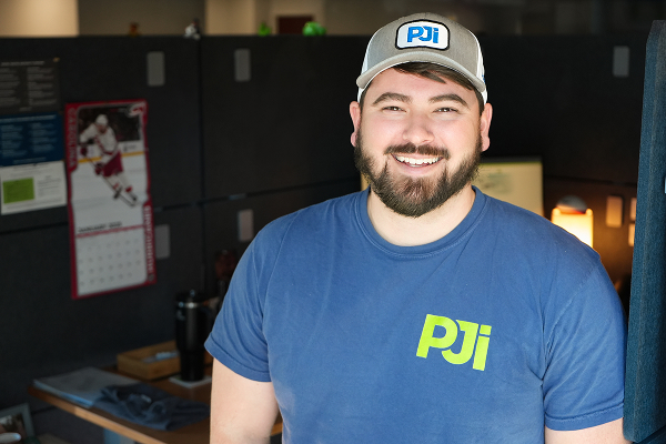 PJi team member smiling in an office workspace wearing a blue PJi shirt and hat with company logo.