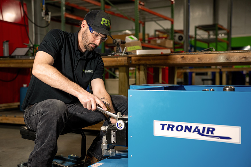 A bearded technician wearing a black shirt and a baseball cap uses a hammer to work on a metal component at a workstation, with a stack of tires and various tools in the foreground.