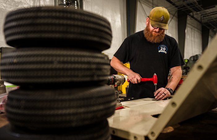  A bearded technician wearing a black shirt and a baseball cap uses a hammer to work on a metal component at a workstation, with a stack of tires and various tools in the foreground.