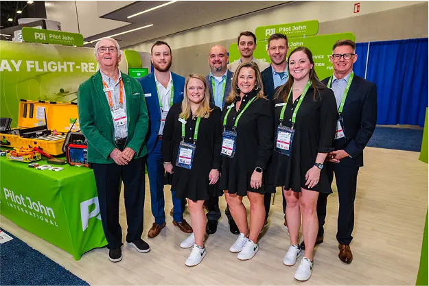 The Pilot John International trade show team posing in front of their booth display with bright green branding and aviation equipment.