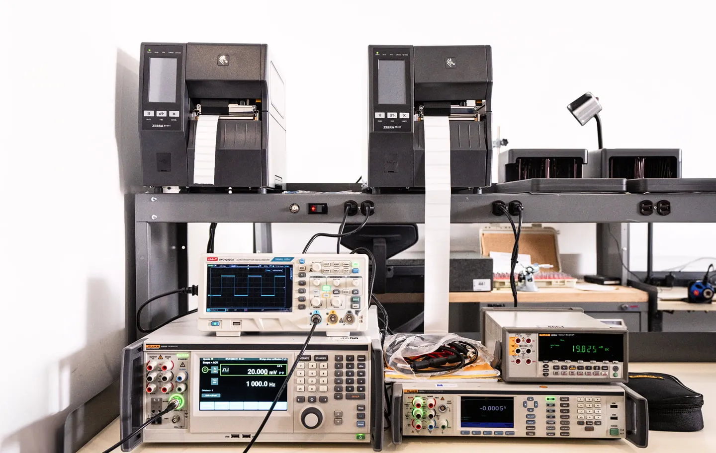 Electronic calibration and measurement instruments on a lab bench with label printers above them.