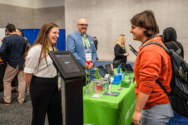 PJi representatives engaging with an attendee at a trade show booth displaying aviation products and promotional materials.