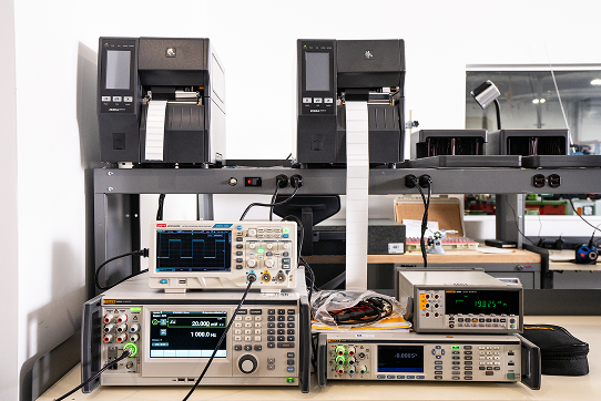 Electronic calibration and measurement instruments on a workbench with label printers above them.