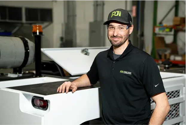 PJi technician wearing safety glasses and a black polo shirt stands next to ground support equipment inside a maintenance facility.