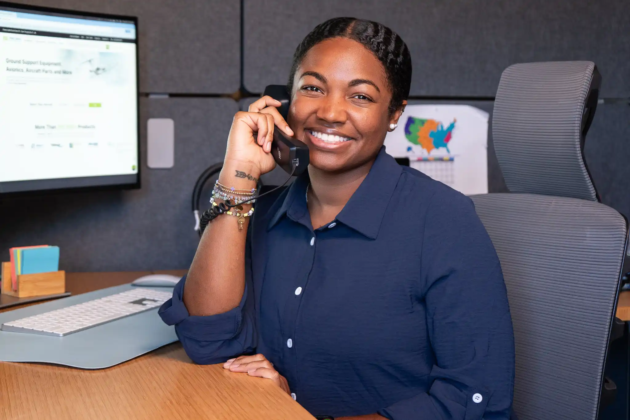 Smiling PJI employee speaking on the phone at her desk, with a computer monitor displaying the Pilot John International website in the background.